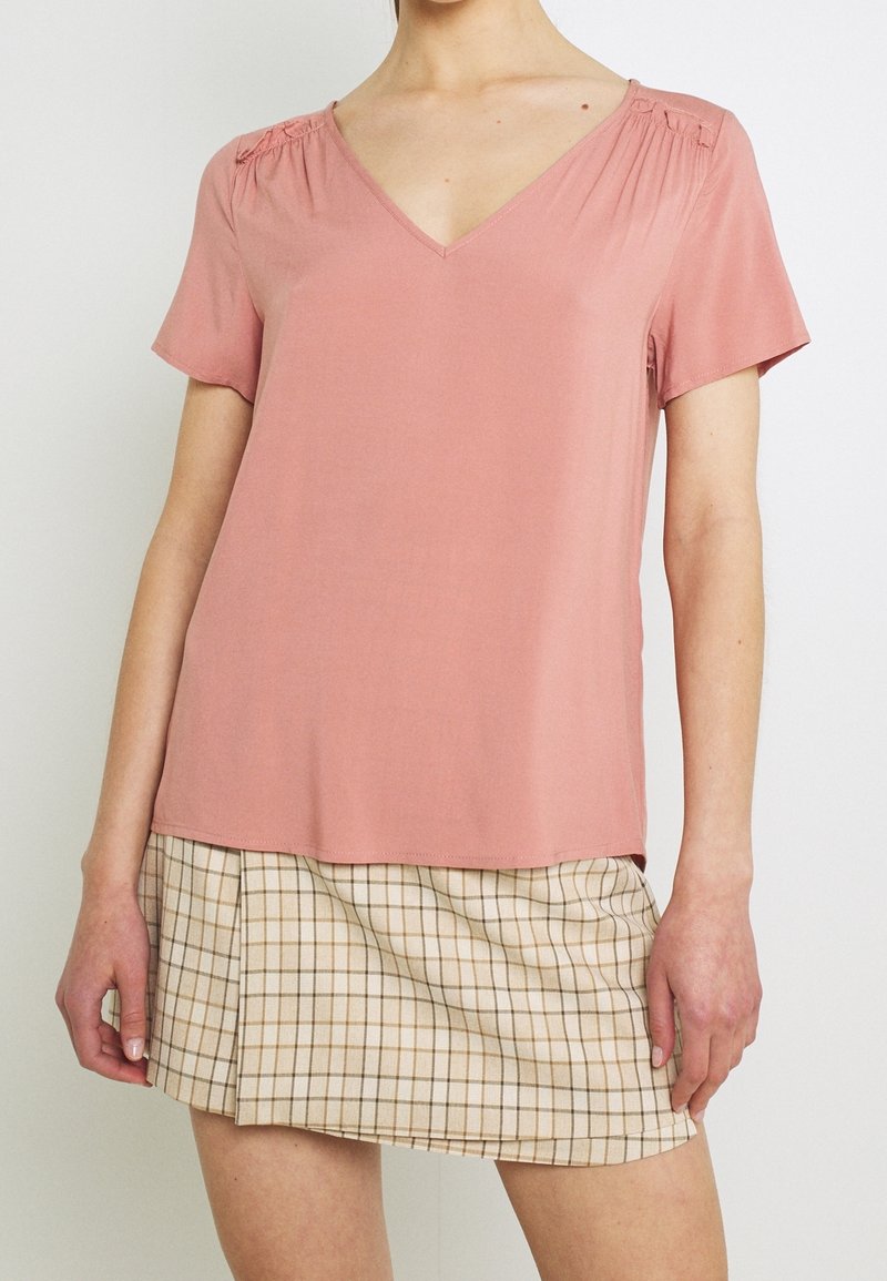 Haut à manches courtes et col en V en tissu rose doux avec détail froncé aux épaules, associé à une mini-jupe beige à carreaux.