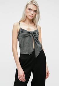 Νεαρή γυναίκα με μακριά ξανθά μαλλιά, φορώντας μια γκρι αμάνικη μπλούζα με κόμπο μπροστά και μαύρο παντελόνι, σε έναν απλό φόντο.