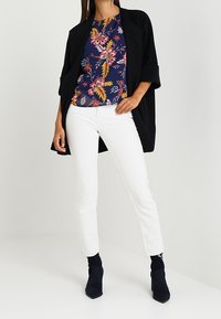 Veste noire à ouverture frontale, haut floral marine, pantalon blanc ajusté et bottines bleues. Le haut présente des motifs floraux colorés.