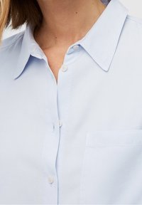 Light blue button-up shirt with a collar, featuring a patch pocket and smooth texture. Buttons are white and rounded.
