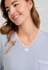 Mujer sonriente que lleva un top azul claro con borde de encaje y un pequeño lazo blanco en el escote, además de un bolsillo con borde de encaje.