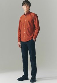 Cămașă de culoare ruginie, cu guler, purtată cu pantaloni negri și pantofi negri. Material neted, croială slim fit și detalii subtile ale nasturilor.