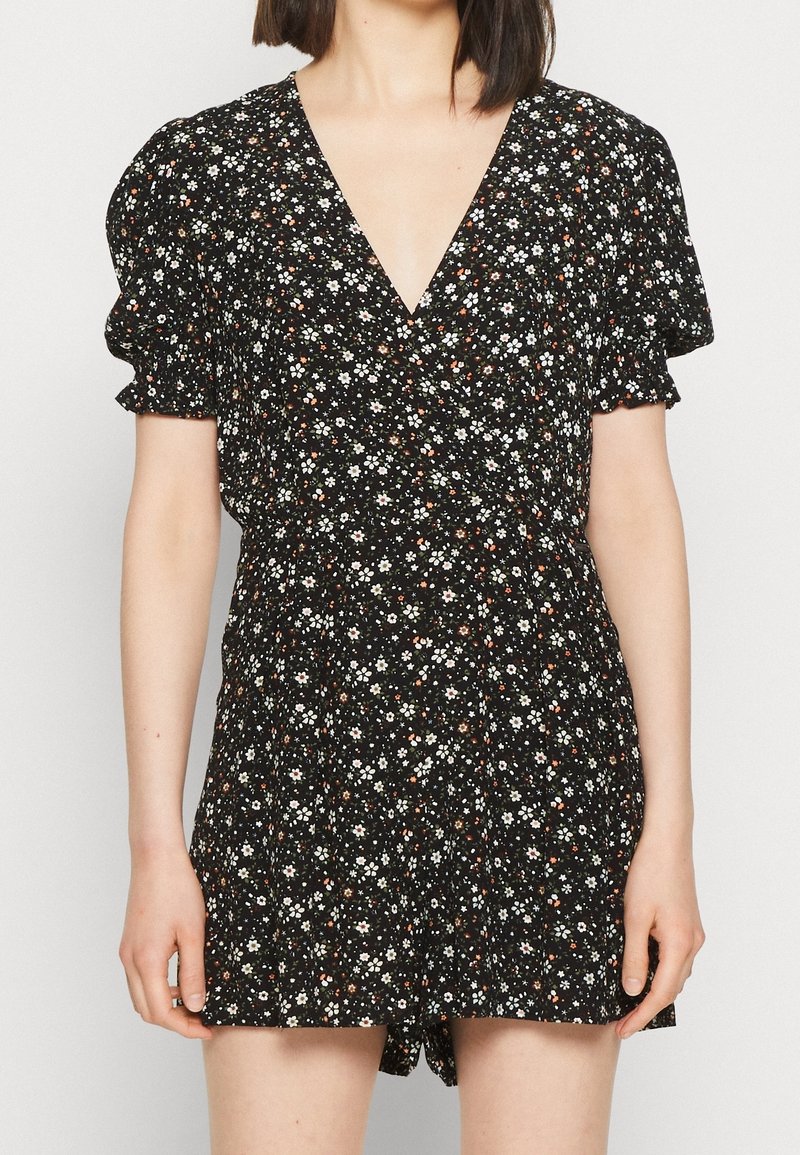 Femme portant une robe courte noire à motif floral avec des manches bouffantes et un profond décolleté en V, debout devant un fond clair uni.