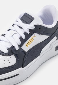Baskets pour homme avec tige en cuir blanc perforé, superpositions synthétiques gris foncé, logo Puma doré et lacets plats blancs. Semelle extérieure en caoutchouc.