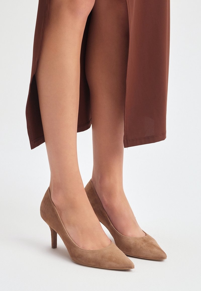Femme portant des talons en daim beige à bout pointu et une jupe marron avec une fente à l'avant, debout sur une surface blanche.