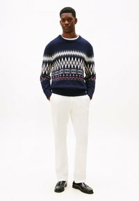 Jersey azul marino con patrones geométricos en blanco y rojo, con puños y dobladillo acanalados, combinado con pantalones blancos y mocasines negros.
