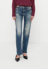 Blugi din denim albastru decolorat, cu croială dreaptă a piciorului, având pete uzate și tiv aspru. Purtat cu pantofi cu toc negri, cu vârf ascuțit.