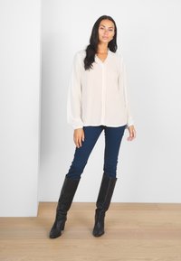 White long-sleeve blouse with a buttoned neckline, paired with dark blue fitted jeans and knee-high black leather boots, standing on light wood floor.