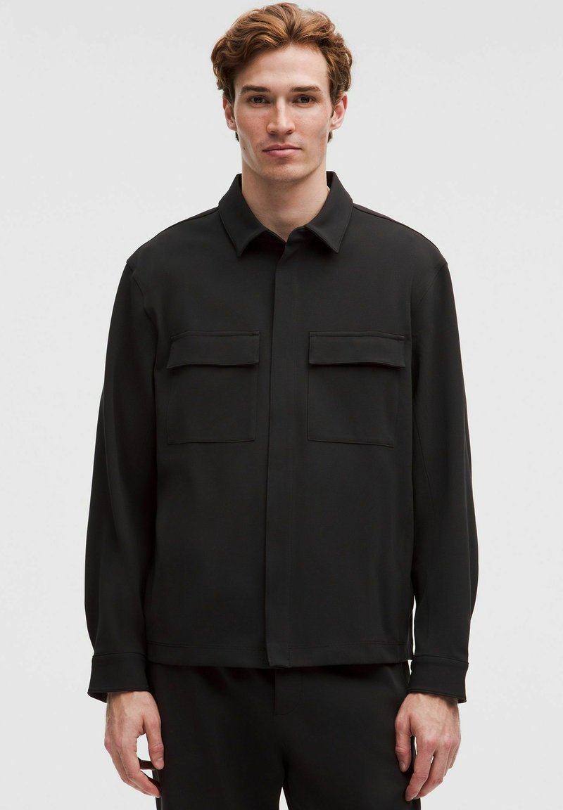 Must müts, mis on valmistatud siledast kangast; sellel on krae, kaks rinna taskut ja pikad varrukad, millel on nööpidega mansetid.