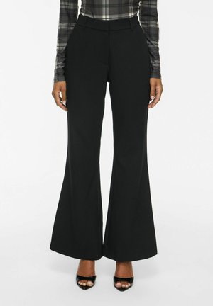 Femme portant un pantalon large noir taille haute, un haut à manches longues à carreaux, et des talons noirs ouverts, debout sur un fond blanc.