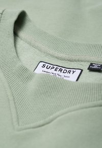 Jasnozielona bluza z prążkowanym kołnierzem i metką z napisem "SUPERDRY" oraz informacjami o lokalizacji. Miękka w dotyku i o casualowym kroju.