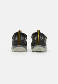 Pari saappaita lapsille musta-harmaa, joissa on keltaiset vetonauhat, nähtynä takaa valkoisella taustalla.