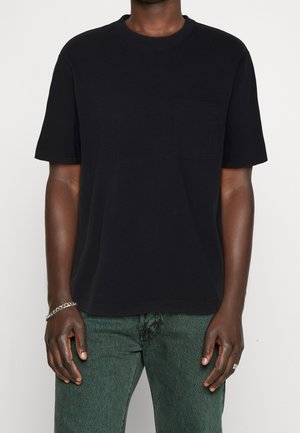 Person iført en enkel sort kortærmet T-shirt med brystlomme og mørkegrønne jeans, stående mod en hvid baggrund.