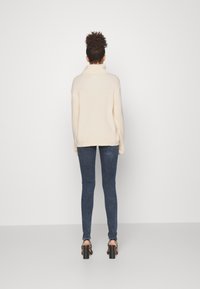 Donna con capelli ricci raccolti in uno chignon, che indossa un maglione a collo alto color crema, jeans skinny scuri e sandali neri con tacco, in piedi davanti a uno sfondo neutro.