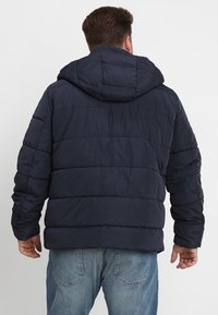 Marineblå pufferjakke med hætte, udstyret med horisontalt quiltet design og elastiske manchetter. Materialet ser blødt og vandafvisende ud.
