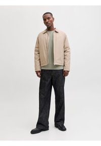 Άνδρας που στέκεται φορώντας μπεζ ζακέτα με φερμουάρ, πράσινο φούτερ, φαρδιά σκουρόχρωμα τζιν και μαύρα παπούτσια, μπροστά σε απλό φωτεινό φόντο.