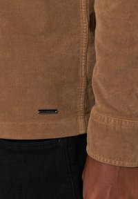 Een bruine corduroy jas met een ribbeltextuur en een metalen label met merk bij de zoom. Draagt over donkere spijkerbroeken met een knoopdetail.