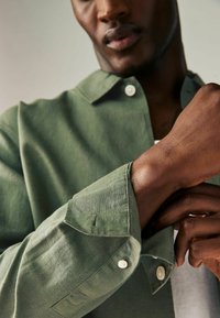 Zeleno laneno srajco z ovratnikom, belimi gumbi in zavihanimi rokavi. Tkanina ima mehko teksturo in rahlo sijaj.