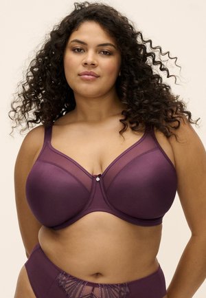 Donna con capelli ricci che indossa un reggiseno viola a copertura totale e mutandine a vita alta abbinate su uno sfondo neutro.