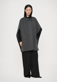 Szara wełniana peleryna o luźnym kroju i zaokrąglonym kształcie, z czarnym zapięciem przy szyi, zestawiona z czarnymi spodnimi o szerokich nogawkach.