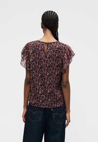 Personne avec des cheveux tressés portant un chemisier noir et rose à fleurs avec des manches à volants et un jean bleu foncé, vue de dos.