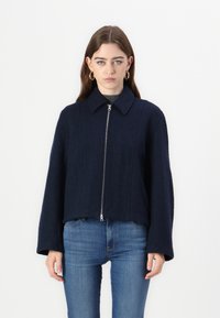 Giacca cropped blu navy realizzata in tessuto strutturato, con chiusura a zip, colletto appuntito e maniche larghe. Indossata sopra un top grigio e jeans.