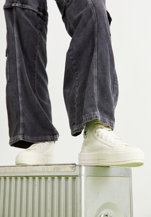Baskets montantes blanches avec une finition texturée, dotées d'une semelle épaisse et d'un patch logo circulaire, associées à un pantalon large gris.