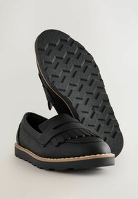 Chaussures synthétiques noires avec des franges, dotées d'une semelle en caoutchouc texturé et d'un contour de couleur claire contrastant.