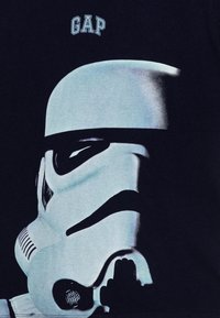 Juodas marškinėliai su Stormtrooper šalmą vaizduojančia grafika mėlynose spalvose, viršuje su "GAP" logotipu. Lygus audinys su drąsiu dizainu ir detalėmis.