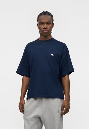 Ung mand med flettede hår iført en mørkeblå Adidas t-shirt og lysegrå Adidas træningsbukser, stående mod en ensfarvet hvid baggrund.
