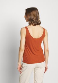 Tank top w kolorze rdzy z dekoltem na plecach, wykonany z prążkowanej tkaniny, z wąskimi ramiączkami i luźnym krojem. W zestawie z jasnymi spodniami.