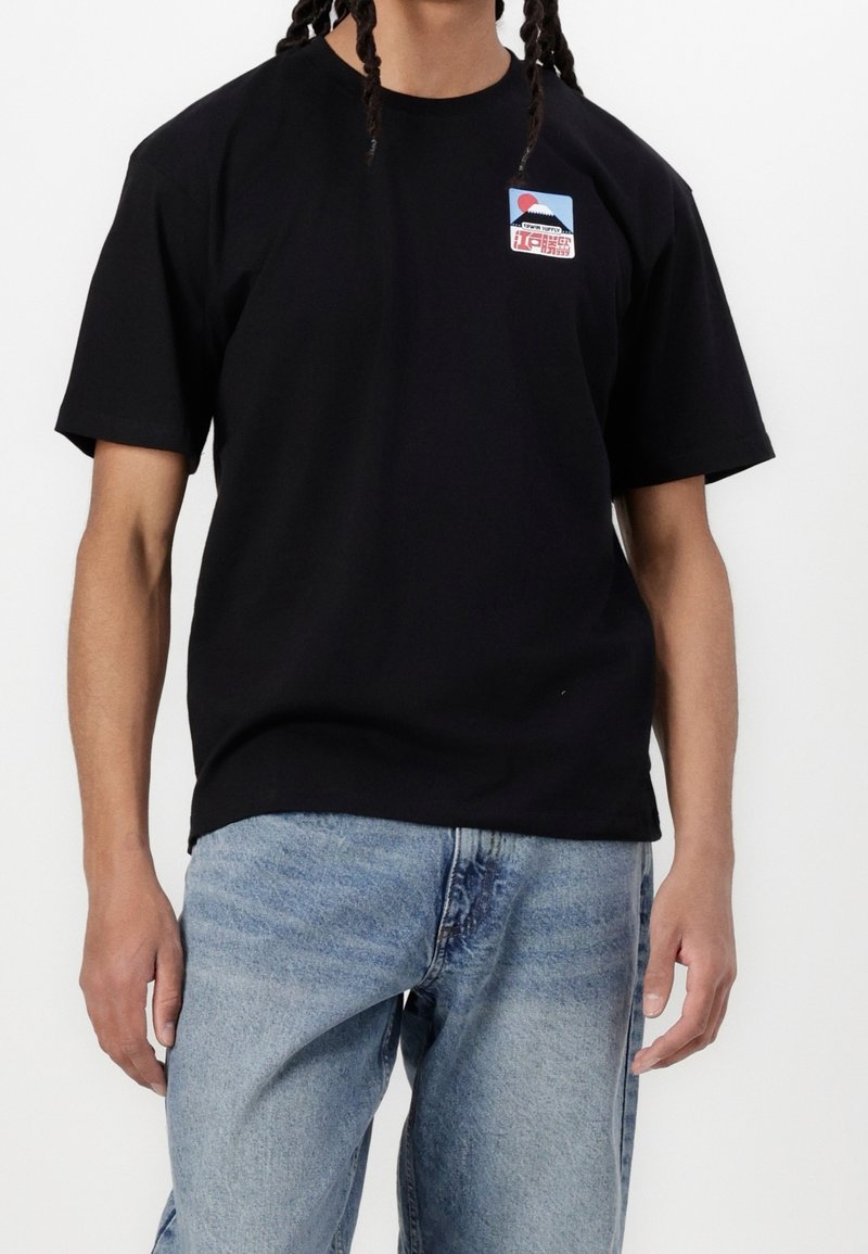 Personne aux cheveux tressés porte un t-shirt noir à manches courtes avec un petit patch coloré sur le côté gauche de la poitrine et un jean bleu clair.