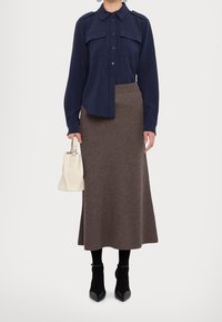 Chemise bleu marine à boutons avec deux poches poitrine, associée à une longue jupe marron côtelée. Le modèle tient un petit sac à main crème et porte des bottines noires.