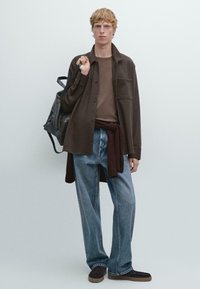 Bruine overhemd met knopen over een bruin shirt, lichtblauwe jeans en donkerbruine instappers. Zwarte rugzak over één schouder.
