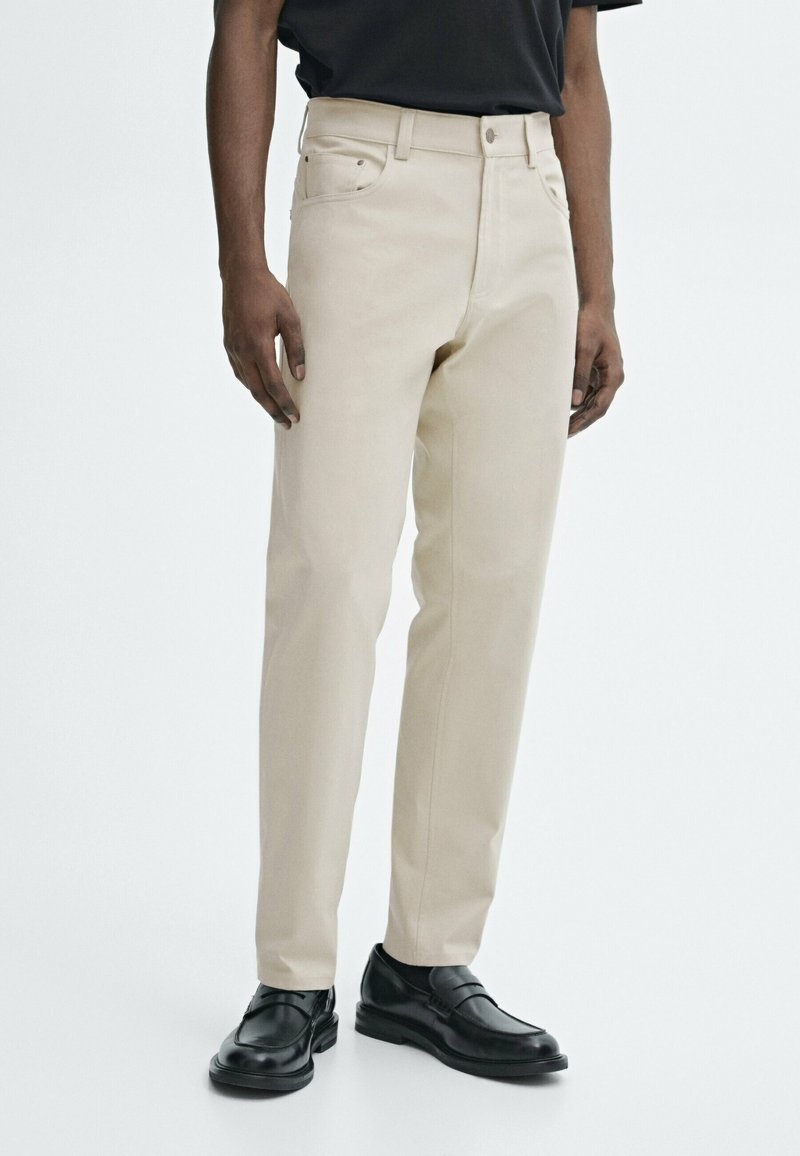 Homme portant un pantalon beige droit avec des poches avant et des mocassins en cuir noir, debout devant un fond blanc.