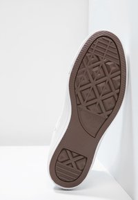 Białe sneakersy z fakturowaną brązową gumową podeszwą z geometrycznymi wzorami i logo, prezentujące gładką cholewkę z detalami w postaci otworów.