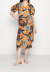 Blouse fleurie colorée à manches ballon, associée à une jupe portefeuille assortie. Présente des motifs audacieux orange, vert et bleu. Portée avec des sandales légères.