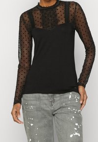 Schwarzes Oberteil mit transparenten Polka-Dot-Langärmeln, Spitzenverzierungen am Ausschnitt und einem taillierten Oberteil. Kombiniert mit distressed grauen Jeans.
