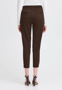 Naine kannab tumepruune lühikesi pükse elastse vöökoha ja mustade kontsadega sandaale, seistes ühtlase valge tausta ees.