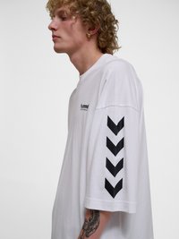 Fiatal személy göndör hajjal, fehér Hummel pólót visel fekete csíkos nyílakkal az ujjon és kis logóval a mellkason, tetoválás látható az alkarján.