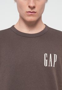 Brown crewneck t-shirt made of cotton, featuring a small white "GAP" logo on the chest, with a smooth, lightweight texture.