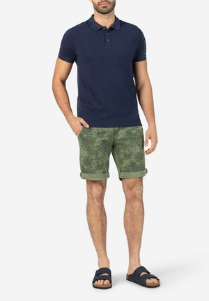 Marineblaues Poloshirt, grüne Camouflage-Shorts und dunkle Sandalen. Das Poloshirt hat einen Kragen und drei Knöpfe. Die Shorts sind an den Säumen umgeschlagen. Lässiges Outfit.