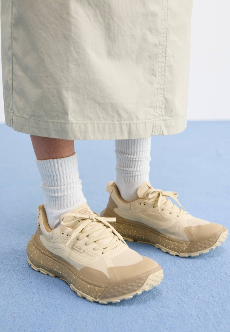 Scarpe da ginnastica beige con tessuto testurizzato, intersuola maculata e lacci bianchi, abbinate a calzini bianchi a coste e una gonna di colore chiaro.