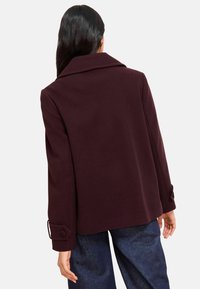 Donna che indossa un blazer di lana viola scuro con polsini abbottonati, vista da dietro, abbinato a jeans blu scuro su uno sfondo uniforme.