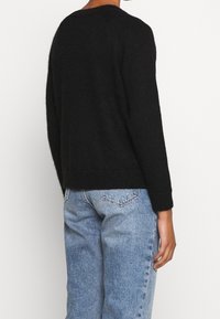 Schwarzer Strickpullover mit lockerer Passform, rundem Halsausschnitt und langen Ärmeln. Trägt dazu hellblaue Jeans, von hinten gesehen.