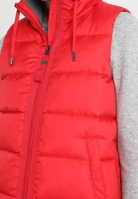 Röd vadderad väst med hög krage, dragkedja fram och snörning i huvan. Har vertikal sömnad och sidofickor med en grå långärmad tröja under.