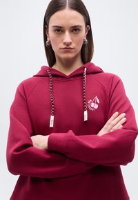 Γυναίκα με ίσια καστανά μαλλιά και ασημένια σκουλαρίκια κρίκους φοράει μπορντό φούτερ με κουκούλα που έχει γραφικό κεράσι και έχει τα χέρια σταυρωμένα μπροστά σε λευκό φόντο.