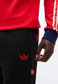 Person wearing black Adidas pants with red logos and a red sweatshirt with white stripes on the sleeve, hand relaxed at side.