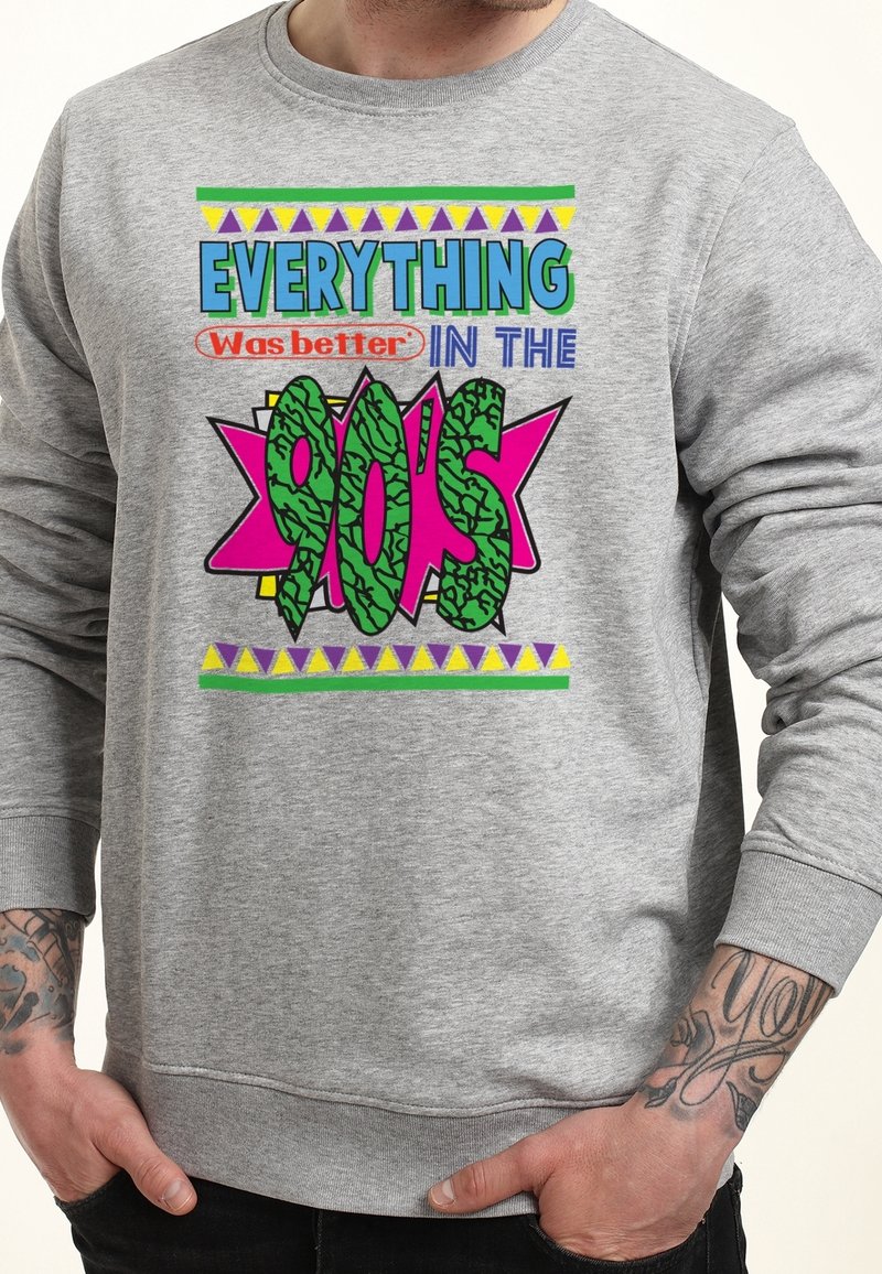 Homme portant un sweat-shirt gris avec un texte coloré "Tout était mieux dans les années 90" et des tatouages visibles sur les deux avant-bras.