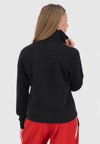 Giacca in pile nera con colletto alto, design aderente e maniche lunghe, abbinata a pantaloni rossi. Il materiale sembra morbido, offrendo calore e comfort.